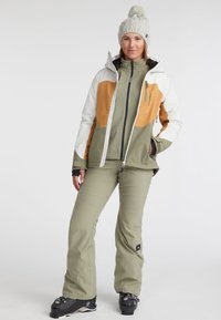 Ski jacket with white, olive, and orange panels, featuring a zip closure and high collar. Paired with olive pants and black ski boots.