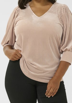 Kobieta w jasnoróżowej prążkowanej aksamitnej bluzce z balonowymi rękawami i czarnych spodniach, trzymająca jedną ręką brzeg bluzki.