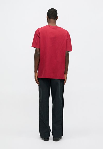 Crvena pamučna majica, opuštenog kroja, okrugli izrez, kratki rukavi; uparen sa širokim crnim hlačama, glatke teksture, bez vidljivih uzoraka.