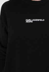 Črna pletena jopica z rebrastim ovratnikom, ki na sprednji strani vsebuje belo vezeno besedilo "KARL LAGERFELD JEANS".