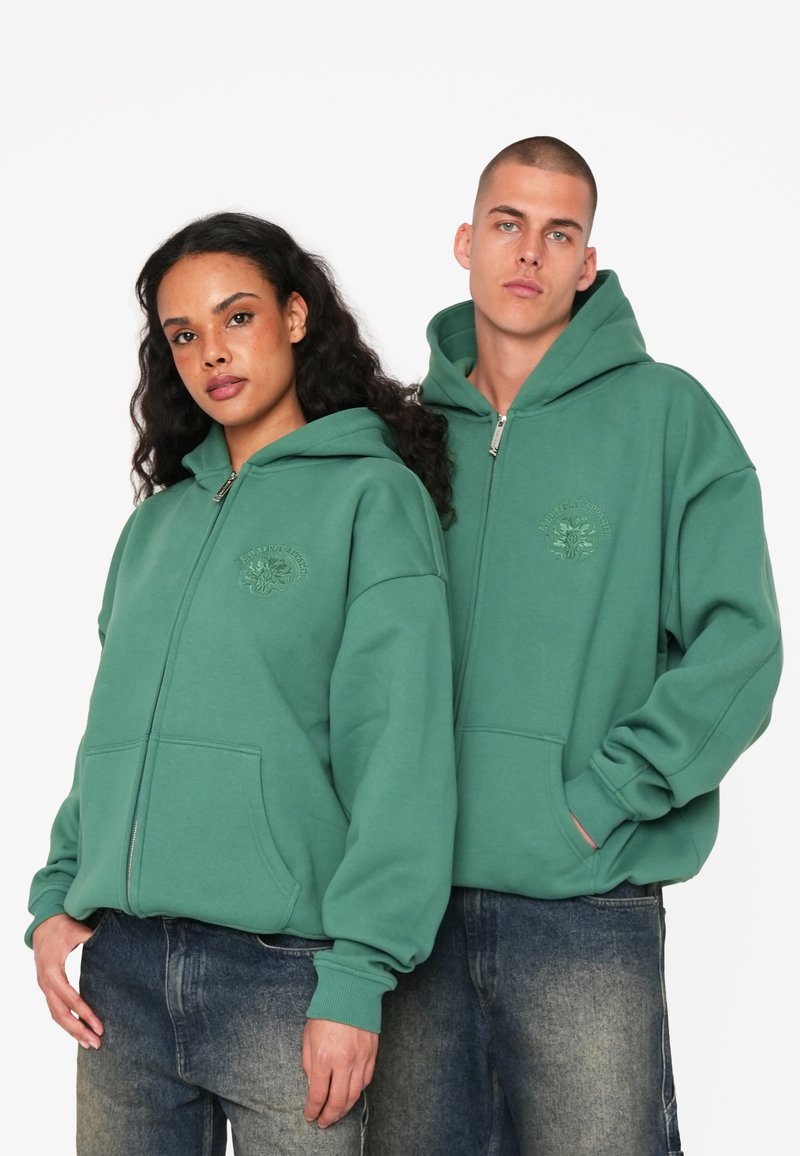 Grüne Reißverschlusskapuzenpullover mit Fronttaschen und einem strukturierten Logo. Beide Modelle tragen lässig sitzende Jeanshosen. Weiche, legere Stofftextur.
