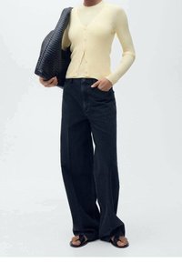 Gerippte gelbe Strickjacke mit Knöpfen, getragen über einem figurbetonten Oberteil, kombiniert mit dunklen, weitem Jeans und schwarzen Sandalen, hält eine strukturierte schwarze Tasche.