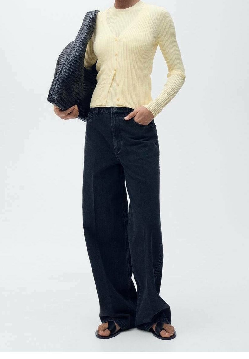 Gerippte gelbe Strickjacke mit Knöpfen, getragen über einem figurbetonten Oberteil, kombiniert mit dunklen, weitem Jeans und schwarzen Sandalen, hält eine strukturierte schwarze Tasche.