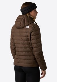 Brązowa kurtka puchowa z poziomym przeszyciem, kapturem i markowym logo na lewym ramieniu. Noszona z czarną, teksturowaną opaską na głowę.
