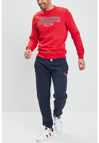Sudadera roja con el texto "CREEKS ORIGINAL Desde 1974", combinada con pantalones de chándal azul marino y zapatillas blancas con detalles en rojo y negro.