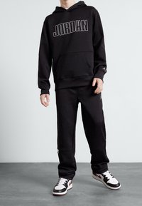 Felpa nera con cappuccio e joggers caratterizzati da sottili motivi geometrici, testo "JORDAN" bianco e tasche con zip. Abbinati a sneakers nere e bianche.