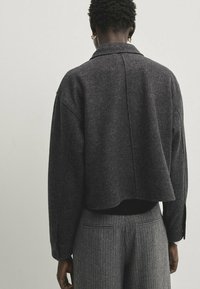 Texturierte, dunkelgraue, kurz geschnittene Jacke mit Kragen und abgesenkten Schultern. Kombiniert mit grau gestreiften Hosen. Keine sichtbaren Verschlüsse oder Verzierungen.