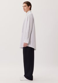 Camicia bianca a righe con una vestibilità rilassata e maniche lunghe, abbinata a pantaloni neri larghi e sneakers bianche, che mostrano un design contemporaneo.