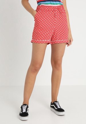 Rote Shorts mit weißen Punkten und weißem Besatz, getragen von einer stehenden Person, kombiniert mit schwarz-weißen Sneakers und einem bunten gestreiften Oberteil.
