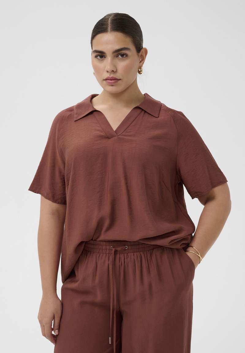 Femme aux cheveux foncés plaqués en arrière, portant un haut marron ample à manches courtes et un pantalon assorti, debout avec une main dans la poche sur fond gris.