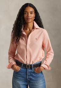 Femme aux longs cheveux bouclés portant une chemise rose clair boutonnée rentrée dans un jean bleu avec une ceinture noire, mains dans les poches.