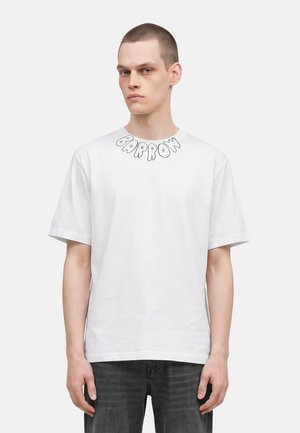 Jeune homme aux cheveux courts portant un T-shirt blanc avec le texte "BARROW" près du col et un jean foncé, debout devant un fond blanc uni.