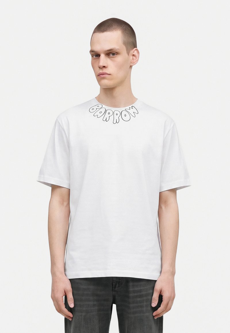 Jeune homme aux cheveux courts portant un T-shirt blanc avec le texte "BARROW" près du col et un jean foncé, debout devant un fond blanc uni.
