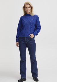 Blått stickat tröja med hög krage, texturerat mönster och ribbad fåll, kombinerad med mörkblå utsvängda jeans och svarta stövlar.