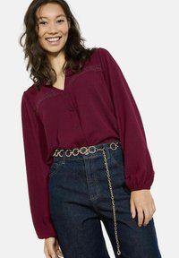 Camicia bordeaux a maniche lunghe con dettagli in pizzo, chiusura con bottoni, abbinata a jeans in denim scuro e una cintura in oro con anelli collegati.