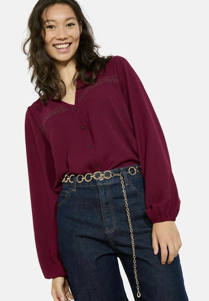 Camicia bordeaux a maniche lunghe con dettagli in pizzo, chiusura con bottoni, abbinata a jeans in denim scuro e una cintura in oro con anelli collegati.