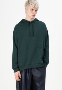 Fiatal személy rövid szőke hajjal, sötétzöld kapucnis pulóvert és fényes fekete nadrágot visel, egyik kezét a zsebében tartva, oldalra néz.