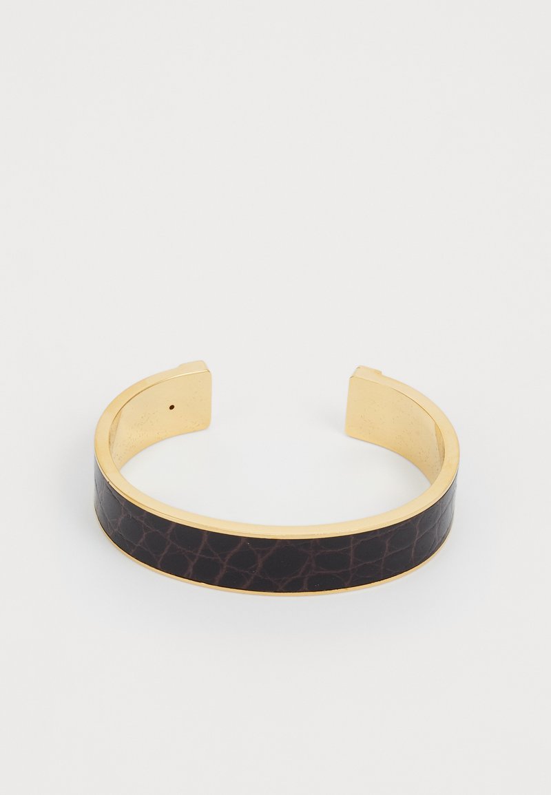 Kuldtooni käevõru, millel on lai tekstureeritud tumepruun nahast riba, mis on kaetud krokodilli mustriga. Avatud otsad mugavaks kandmiseks.