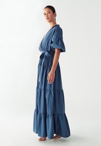 Maxi šaty z tmavě modré tkaniny s nabíranými rukávy, páskem na zavázání v pase a volánky na lemu. Hladký povrch a lehký design.