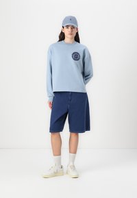Helles blaues Rundhals-Sweatshirt mit navyfarbener Grafik, kombiniert mit dunkelblauen Denim-Shorts und weißen Sneakers mit gelben Sohlen.