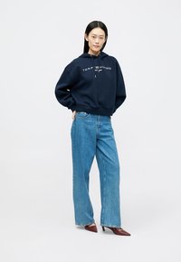 Σκούρο μπλε φούτερ με λευκό λογότυπο, συνδυασμένο με ανοιχτό μπλε παντελόνι με φαρδιά κοψίματα. Το μοντέλο φοράει μπορντό γόβες, στέκοντας μπροστά σε έναν απλό φόντο.