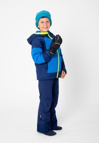 Blauwe en groene winterjas met capuchon, felgele ritsen, donkerblauwe broek en zwarte handschoenen. De outfit omvat een gebreide muts met een pompon.