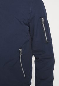 Granatowa kurtka o gładkiej teksturze, z wyraźnymi zamykanymi na zamek kieszeniami na ramionach i z boku oraz ze smukłym fasonem.