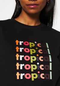 Mujer vestida con una camiseta negra que tiene cinco palabras "tropicales" apiladas en diferentes colores brillantes en el pecho.