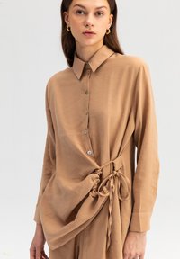 Camicia button-up beige con una tessitura morbida, caratterizzata da maniche lunghe, un dettaglio a nodo sul lato e un design classico del colletto.
