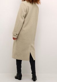 Beżowy płaszcz z mieszanki wełny o teksturowanym wykończeniu, z długimi rękawami, zapinanymi na guziki mankietami oraz rozcięciem z tyłu. Połączony z ciemnymi jeansami i botkami za kostkę.