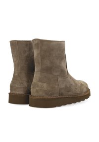 Un par de botas de tobillo de ante marrón con suelas de goma planas, vistas desde atrás y en ángulo hacia la derecha sobre un fondo blanco.