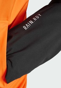 Vandtæt jakke med en orange krop og sorte ærmer. Har "RAIN.RDY" logo i hvidt på det sorte stof. Glat tekstur.