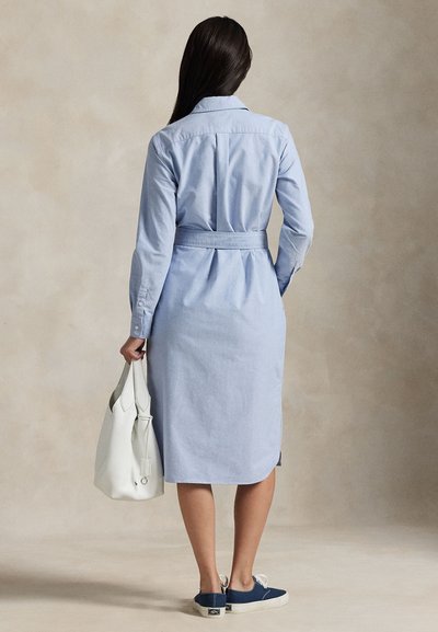Moteris su ilgais tamsiais plaukais vilki šviesiai mėlyną su diržu marškinių suknelę, laiko baltą rankinę ir dėvi tamsiai mėlynas sportbačius, stovi prieš neutralią foną.