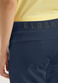 Navy blue athletic pants featuring a wide elastic waistband with embossed lettering and a small brand tag on the side. Soft stretch fabric.