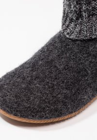 Grau wollene Hausschuhe mit einer dicken, flauschigen Textur; die weiche Sohle hat einen braunen Gummi-Besatz, und der gestrickte Bund hat ein dunkleres graues Muster an der Oberseite.
