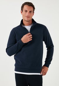 Pullover blu navy con colletto a mezzo zip, realizzato in morbido tessuto. Presenta maniche lunghe e una tasca frontale piatta. Texture liscia con polsini a coste.