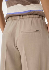 Primo piano di pantaloni beige a vita alta con tasche a filetto sul retro e un piccolo etichetta in metallo del marchio, abbinati a un top a righe marrone e viola con coste.