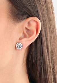 Primer plano de la oreja de una mujer con cabello castaño lacio, que lleva un pendiente de plata redondo con múltiples pequeños cristales transparentes.
