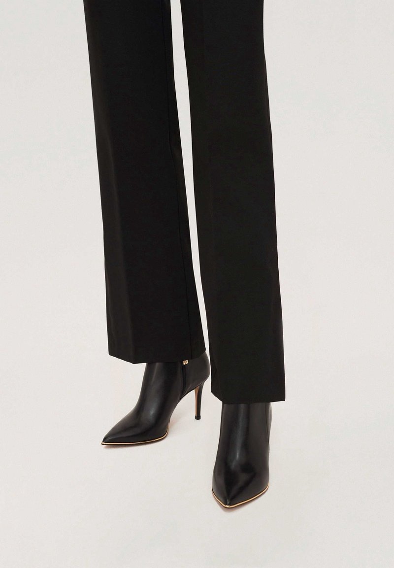Bottes noires en cuir à bout pointu avec talons fins portées sous un pantalon noir droit sur un fond clair.