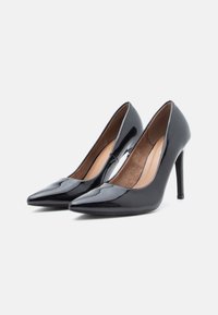 O pereche de pantofi cu toc înalt din piele patentă neagră, cu vârful ascuțit, pentru femei, pe un fundal alb.