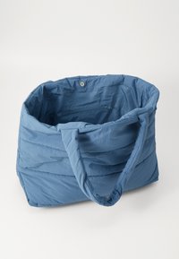 Sac fourre-tout en bleu rembourré avec deux poignées, comportant une poche intérieure zippée et un tissu doux et texturé. Forme arrondie avec des accents cousus.