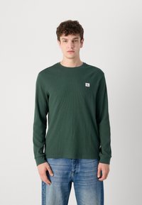 Olijfgroene T-shirt met lange mouwen, gemaakt van een gestructureerde stof en met een ronde halslijn, voorzien van een klein logopatch op de borst. Gecombineerd met blauwe jeans.