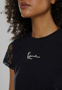 Svart t-skjorte med rund hals og korte blondeærmer. Brodert hvit signaturdetalj på brystet. Myk tekstur.