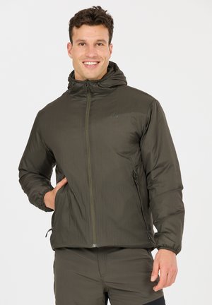 Mies hymyilee, pukeutuneena oliivinvihreään vetoketjulliseen huppariin ja siihen sopiviin housuihin, käsi takin taskussa.