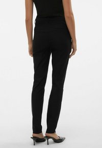 Pantalones negros ajustados con una textura suave, con frente plano y bolsillos traseros, combinados con zuecos negros de tacón, vistos desde atrás.