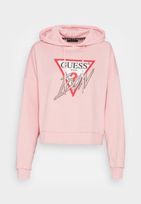 Jasnoróżowa krótka bluza z kapturem ze sznurkami, z czerwoną trójkątną logo oraz napisem "GUESS" z "Jeans" w błyszczącej czcionce z przodu.
