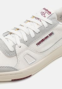 Vita och grå sneakers i läder och mocka, med snörning, en texturerad design och Reebok-logotyp på sidan.