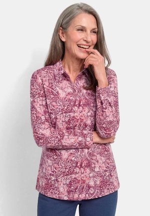Smiling gray-haired woman wearing a pink patterned long-sleeve shirt and blue pants, standing with one hand near her mouth.