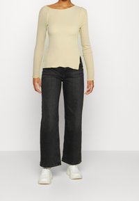 Beige pitkähihaine paita, jossa on keltaiset ompeleet, yhdistetty tummanharmaisiin leveälahkeisiin farkkuihin ja valkoisiin tennoihin, esittelee rennon asukokonaisuuden.