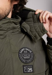 Chaqueta acolchada verde oliva con un diseño de acolchados, que presenta una capucha negra desmontable y parches de logo de goma en el pecho y la manga.